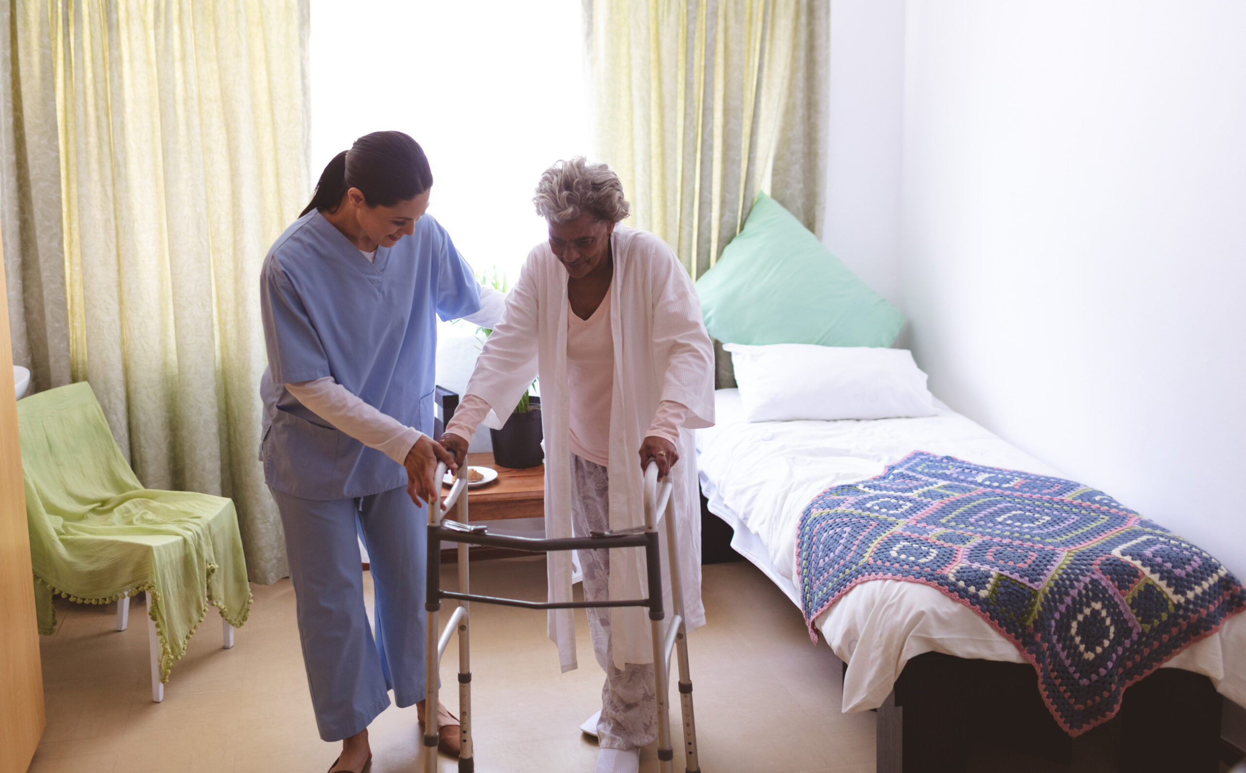 Front view of mixed race female nurse helping senior  mixed race female patient to stand with walker at nursing home